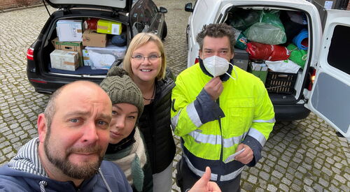 Zwei Autus von hinten mit offenem Kofferraum. Voll beladen mit Kisten und Säcken. Selfie von 2 Männern und 2 Frauen die hinter den Autos stehen.