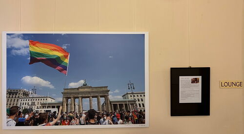 Ausstellungsraum mit gerahmtem Foto vom Brandenburger Tor in Berlin, viele Menschen und einer schwenkt die Regenbogenflagge. an der Wand und kleinem Begleittext daneben.