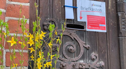 Robuste Holztür mit Briefkastenschlitz, daneben ein Hinweiszettel mit Pfeil zum Briefkasten.