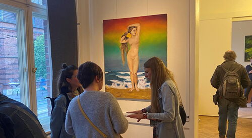 Ausstellungsraum. Drei Frauen stehen vor einem Bild an der Wand. Das Bild zeigt eine nackte Frau mit langen Haaren am Strand stehend. Die Hintergrund ist Regenbogen-farbig.