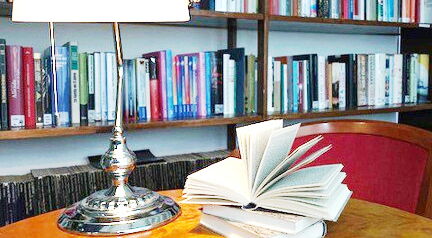 Auf einem Tisch eine Leselampe und 3 Bücher das oberstte ist aufgeschlagen. Dahinter Bücherregale.