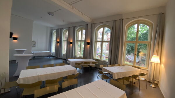Im Raum stehen vier Tische mit Tischdecken und je 8 Stühlen um den Tisch. Im Hintergrund stehen 2 Stehtische