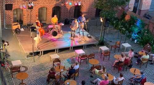 Innenhof mit kleinen runden Tischen und Stühlen, einer Bühne mit Band und Blick von oben auf die Szene. Wenige Zuschauende sitzen an den Tischen.