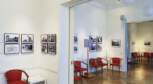 Ausstellungsraum mit Bildern an den Wänden, roten Stühlen und offenem Durchgang zu weiteren Räumen.