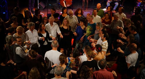 Viele Menschen in einer Halle, die sich in Kreisen an den Händen halten und gemeinsam Tanzen. Oden am Bildrand Bühne mit Band.