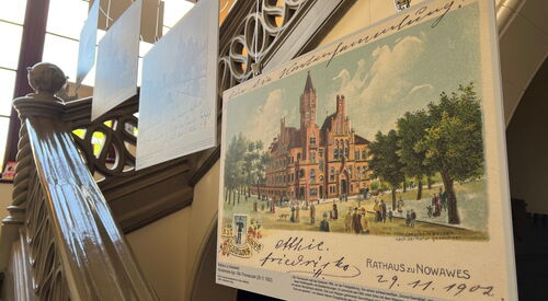 Ausstellungstafeln in einem Treppenhaus. Ausstellungstafel mit historischem Bild des Rathauses Nowawes mit Bäumen davor, gestaltet wie ein Postkartenmotiv, darunter Bildbeschreibungstext.