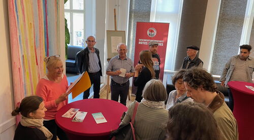 Großer Raum, feierlicher Anlass. Zwei rote Stehtische. Links ein großes Bild an der Wand, davor steht eine blonde Frau und hält einen Hefter und lächelt. Gäste im Raum verteilt.