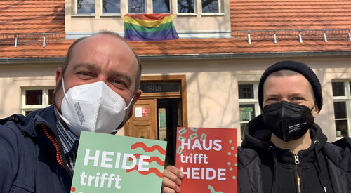 Im Vordergrund zwei Männer mit Maske sie halten jeder ein Schild in der Hand Text: "Heide trifft Haus"  An dem Haus dahinter ist eine Regenbogenflagge.