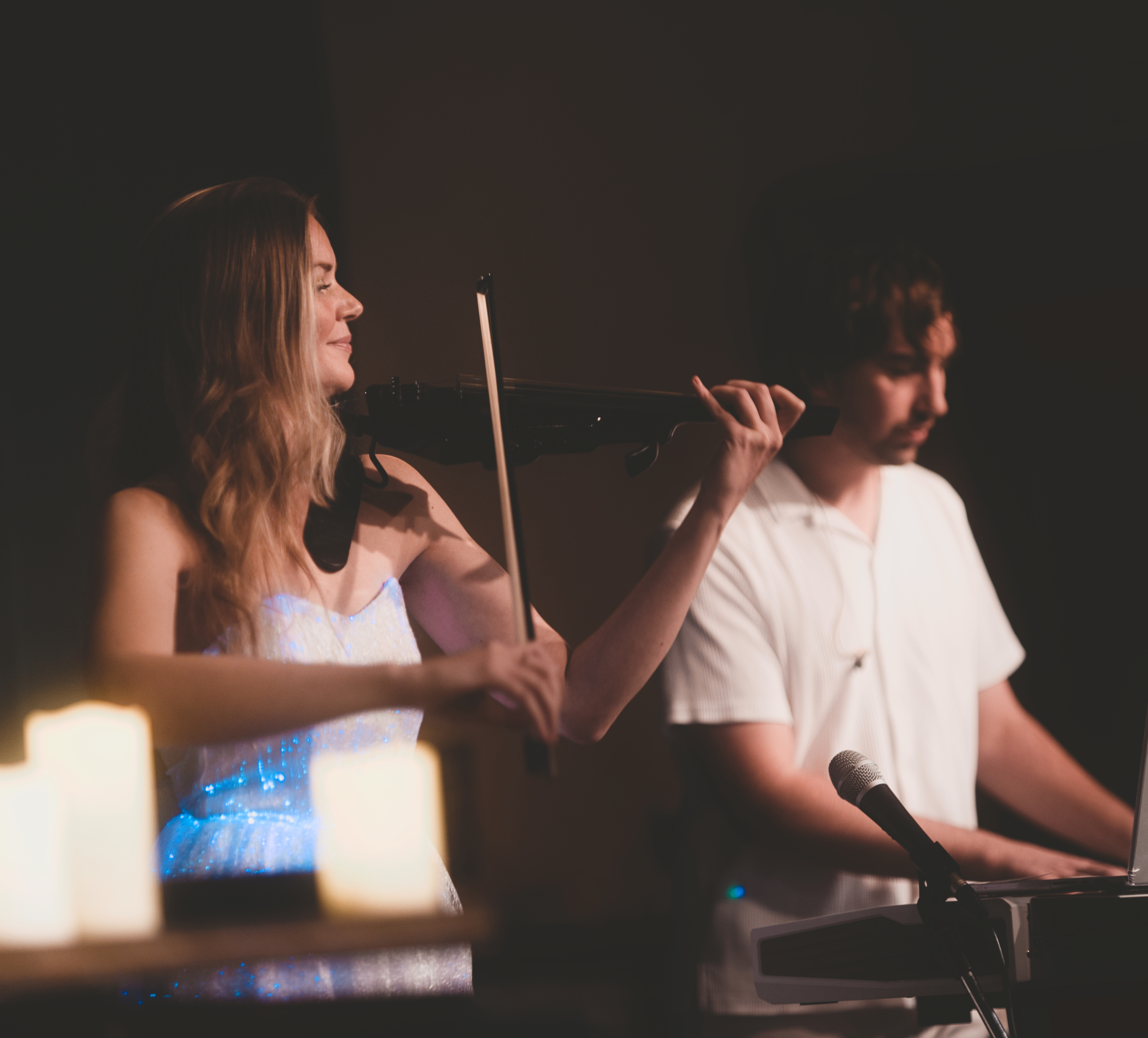 Frau in weißem Glizzerkleid spielt Geige, Mann mit weißem lockeren Hemd neben ihr am Keyboard.