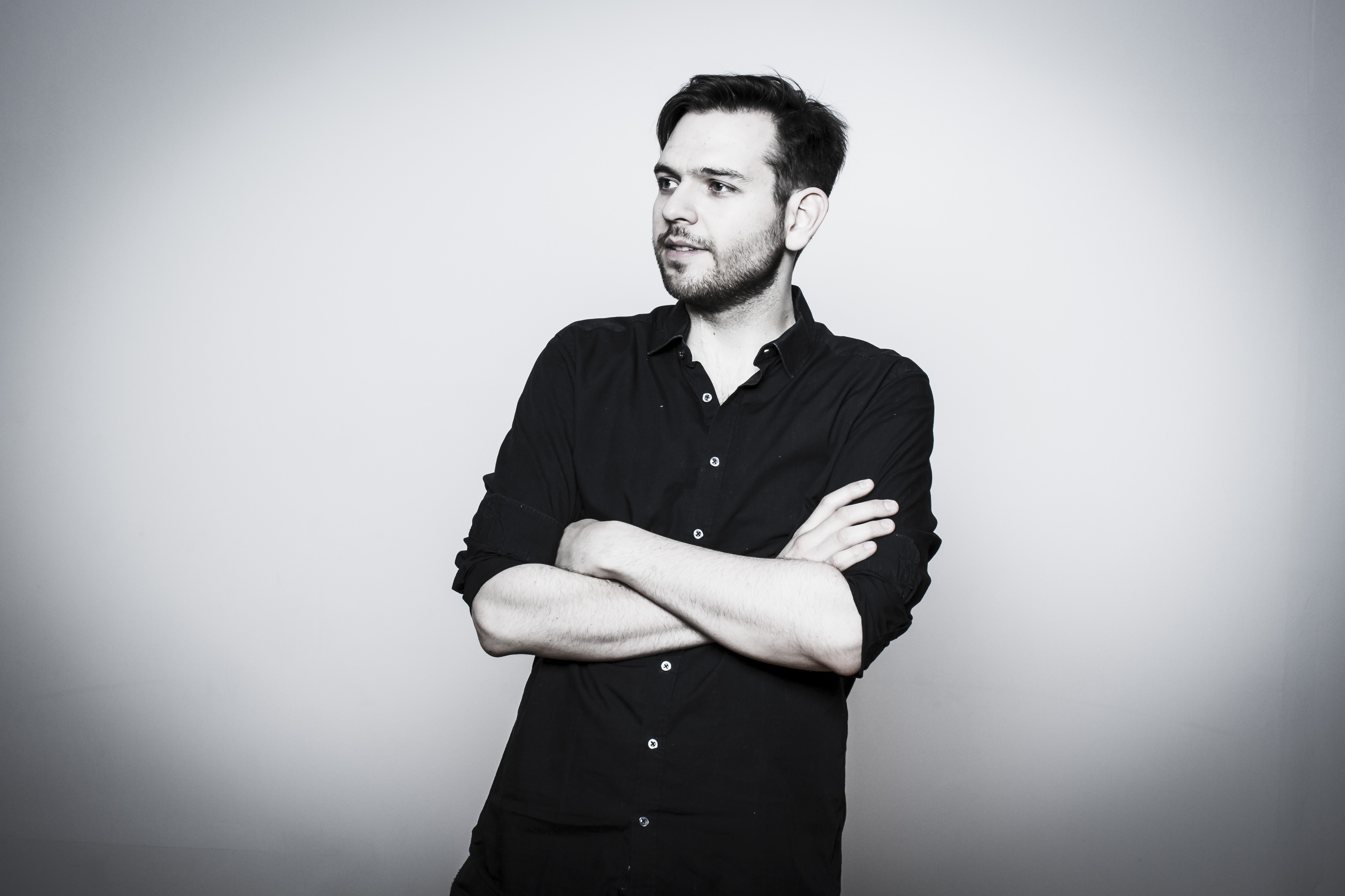 Schwarz-Weiß Foto: Junger Mann im schwarzen Hemd, dunklen Haaren und Bart. Arme vor der Brust verstränkt. Lässig.