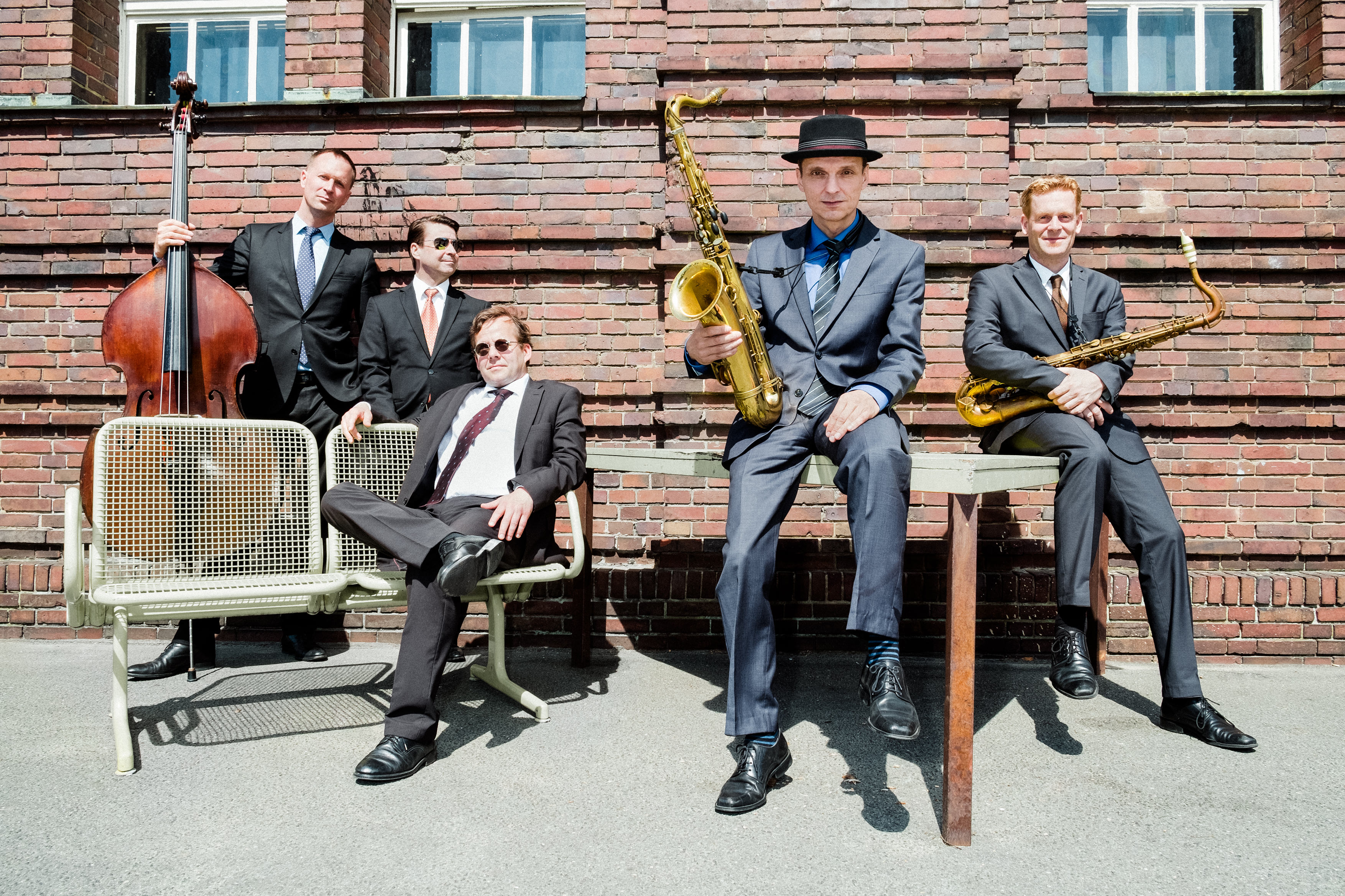 Fünf Männer in grauen und schwarzen Anzügen mit Krawatte stehen und sitzen locker vor einem Gebäude und halten Musikinstrumente.